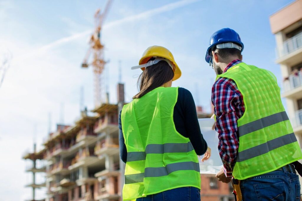 A man and a woman standing side by side at a construction site. They're wearing safety vests and hard hats.